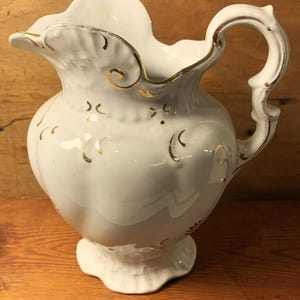 May include: White ceramic jug with a ruffled rim and ornate handle. The jug is decorated with gold accents and a floral design. The base is scalloped. A decorative piece for serving beverages or displaying flowers.