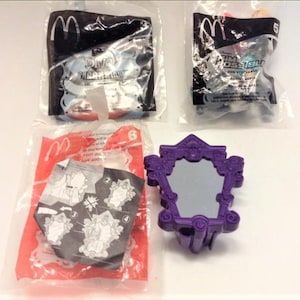 May include: Three unopened plastic bags containing McDonald's Happy Meal toys. The toys are from the Disney movie "The Princess and the Frog". The toys are a purple mirror, a blue and white frog, and a green and white frog. The bags are labelled with the McDonald's logo and the movie title.