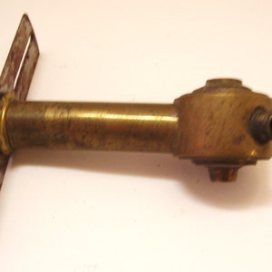 May include: A brass pipe fitting with a flat metal plate attached to one end. The pipe fitting has a round, decorative end with a small hole. The metal plate is rusty.