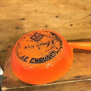 Peut inclure: Une poêle Le Creuset orange vif avec une longue poignée. Le fond de la poêle porte le nom de la marque et l'inscription "Made in France". La poêle a un diamètre de 20 cm.