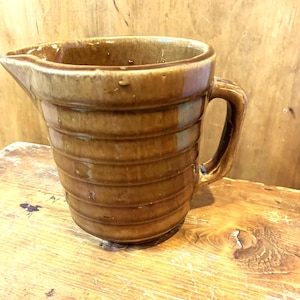 May include: A brown ceramic jug with a wide mouth and a handle. The jug has a ribbed design and is glazed in a brown colour.
