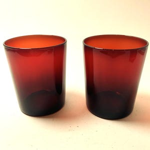 Peut inclure: Deux verres à boire teintés en rouge. Les verres présentent un effet dégradé, passant d'un rouge plus clair en haut à une teinte plus foncée à la base. Les verres sont cylindriques avec un dessus légèrement évasé.