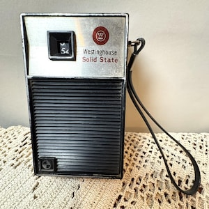 May include: A vintage Westinghouse Solid State portable radio featuring a rectangular black plastic body with a ribbed speaker grille, a silver-toned top panel with a black tuning dial displaying the number 54, and a black wrist strap attached.