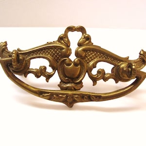 Peut inclure: Une poignée de tiroir en métal doré avec un design orné. La poignée a une forme incurvée et présente des détails complexes, y compris des volutes et des motifs floraux.
