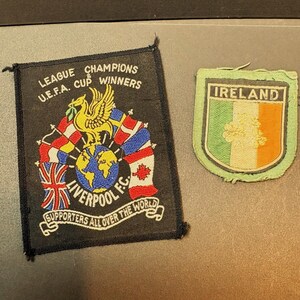 May include: A rectangular black Liverpool F.C. patch featuring a yellow Liver bird, a globe, and international flags with the text LEAGUE CHAMPIONS U.E.F.A. CUP WINNERS LIVERPOOL F.C. SUPPORTERS ALL OVER THE WORLD, alongside a shield-shaped Ireland flag patch.