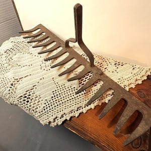 May include: An old, rusty metal garden rake with a long handle and curved tines. The rake is placed on a wooden surface and a cream-coloured, crocheted lace cloth. The rake's design suggests it is for gardening.