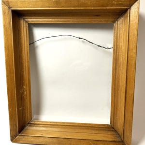 May include: A rectangular, wooden picture frame with a light brown finish. The frame has a grooved design and a wire for hanging. The interior is empty, revealing a white background.