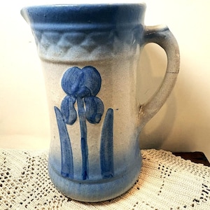 Peut inclure: Un pichet en grès au corps gris clair avec un vernis dégradé bleu en haut et en bas. L'avant présente un motif de fleur d'iris bleu en relief, tandis que le bord supérieur est orné d'un motif en losange texturé et d'une anse latérale.