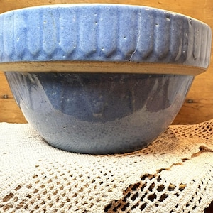 Peut inclure: Un grand saladier en céramique bleu avec un extérieur texturé. Le bol a un large bord avec des rainures verticales et de petits détails en pointillés. Le bol repose sur un napperon en crochet crème, avec un fond en bois.