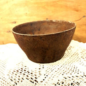 May include: A weathered, brown metal bowl with a flared design. The bowl has a rustic, aged look, possibly vintage or antique. It rests on a white, open-weave lace doily, with a wooden backdrop.