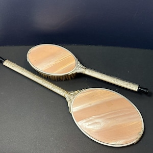 May include: Two oval-shaped hand mirrors with long, silver-toned metal handles featuring engraved geometric patterns. The back of each mirror has a smooth, light peach-coloured inlay with a subtle pearlescent sheen and a black tip on the handle.