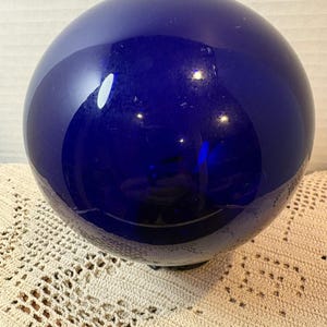 May include: A smooth, spherical object made of deep cobalt blue glass. The surface is reflective and glossy, showing light highlights and a dark, circular base at the bottom. The sphere is solid in colour and has a uniform, rounded shape throughout.