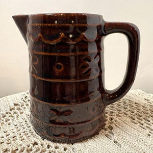 May include: A dark brown ceramic jug with a glossy finish, featuring a cylindrical body, a side handle, and a pouring spout. The exterior surface is decorated with horizontal bands of embossed geometric patterns and scalloped textures.