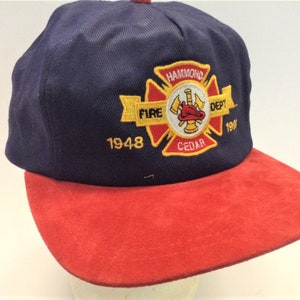 May include: A blue and red baseball cap with an embroidered patch featuring a red cross with a fire helmet, axe, and hose. The patch reads "Hammond Fire Dept. Cedar 1948 199?"