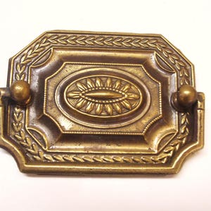 May include: A brass cabinet pull with a decorative design featuring a central oval with a floral pattern. The pull has two rounded knobs on either side.