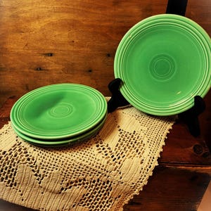 May include: A collection of green ceramic plates. Three plates are stacked on a beige lace doily, while another is displayed on a black stand. The plates have concentric ring designs.