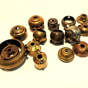May include: A selection of antique brass and metal lamp components, including caps, bases, and decorative elements. The pieces display diverse shapes and sizes, with signs of age and wear, presenting a warm, vintage aesthetic. The components are arranged on a white surface.