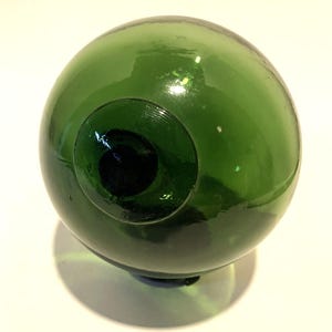 以下が含まれることがあります： 中央に暗い穴のある緑色のガラス球。球体は小さく丸い台座の上にあります。ガラスは光を反射する光沢のある仕上がりです。このオブジェクトはおそらく装飾品またはガラス製のフロートです。