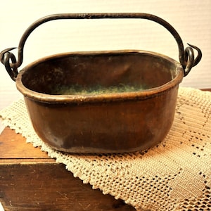 May include: An oval-shaped copper vessel with a dark, weathered patina and a green oxidised interior. It features a thick, arched iron handle attached to the sides with curled metal loops, showing signs of age and wear on the metal surfaces.