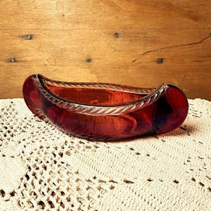 May include: A deep red glass bowl shaped like a canoe, featuring a translucent body and a textured, rope-like pattern along the upper rim. The vessel has a smooth, curved base and a wide, open top, showcasing a rich, dark crimson colour throughout.