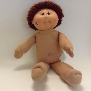 May include: A soft-bodied doll with a round, peach-toned fabric face, brown yarn hair, and brown eyes. The doll features a small blue signature mark on the right cheek, a navel indentation, and is wearing a white fabric nappy on its lower torso.