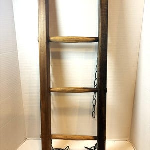 May include: A vertical wooden ladder with two dark brown rectangular side rails and three lighter brown, rounded horizontal rungs. A thin black metal chain is attached to the side and bottom of the ladder structure, hanging loosely from the frame.