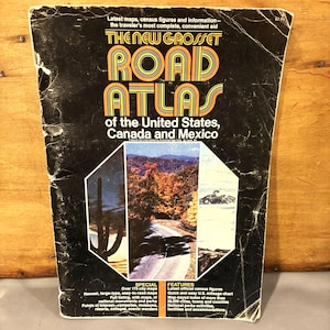 以下が含まれることがあります： 「The New Grosset Road Atlas of the United States, Canada and Mexico」と題されたヴィンテージのロードアトラス。表紙は黒色で、オレンジ色と黄色の文字が書かれています。風景の写真や、都市地図や国勢調査の数字などの情報が掲載されています。
