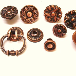 Peut inclure: Assortiment de dix boutons et poignées de placard en cuivre avec des motifs ornés. Les boutons sont ronds et présentent divers motifs floraux et géométriques. Les poignées ont une forme de bague avec un centre décoratif.