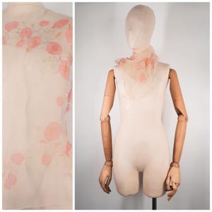 Op de afbeelding: Een crèmekleurige, transparante sjaal met een delicaat roze bloemenpatroon. De lichte stof is gedrapeerd rond een mannequin, wat de lichte en luchtige textuur benadrukt. Het bloemenpatroon herhaalt zich over de stof.