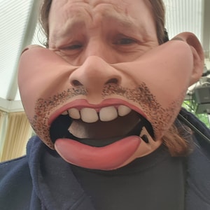 Puede incluir: Una persona lleva una máscara de títere grande, exagerada, marrón y rosa con la boca muy abierta, mostrando dientes blancos y una lengua negra. La máscara tiene un bigote y una barba marrones.