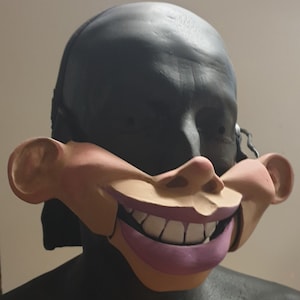 May include: A novelty mask featuring a large, exaggerated smiling face with prominent teeth and pink lips. The mask has a flesh-toned face with large ears and is attached to a black head covering.