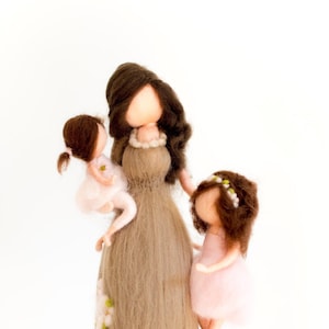 May include: A felt doll representing a mother with two daughters. The mother is wearing a brown dress and has long brown hair. The daughters are wearing pink dresses and have brown hair. The doll is handmade and has a whimsical, handcrafted look.