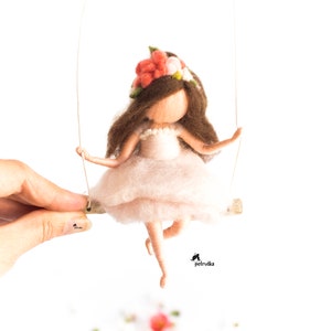 May include: A felt doll with brown hair wearing a pink dress and a flower crown. The doll is on a swing made of wire and wood.