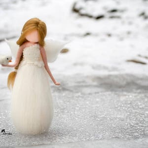 May include: A white felt doll with long brown hair wearing a white dress and holding a white bird. The doll is standing on a snowy surface.