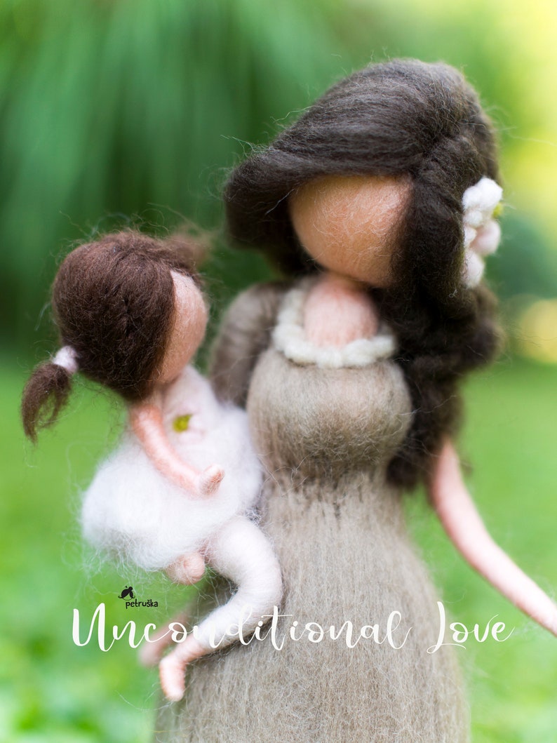 May include: A felt doll of a mother holding a smaller felt doll of a child. The mother doll is wearing a brown dress and has long brown hair. The child doll is wearing a white dress and has brown hair. The dolls are standing on a green grassy surface. The text "Unconditional Love" is written below the dolls.