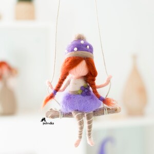 Peut inclure: Une poupée en feutre violette aux cheveux roux portant un chapeau violet et blanc est assise sur une balançoire en branche de bois. La poupée porte une robe violette et des bas blancs.