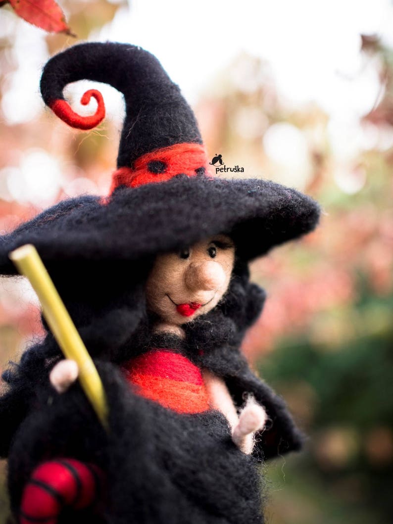 May include: A felt witch doll with a black hat, red and black dress, and a yellow broom. The doll has a friendly face with red lips and a black nose.