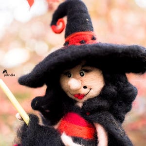 May include: A felt witch doll with a black hat and red and black dress. The doll is holding a yellow stick.