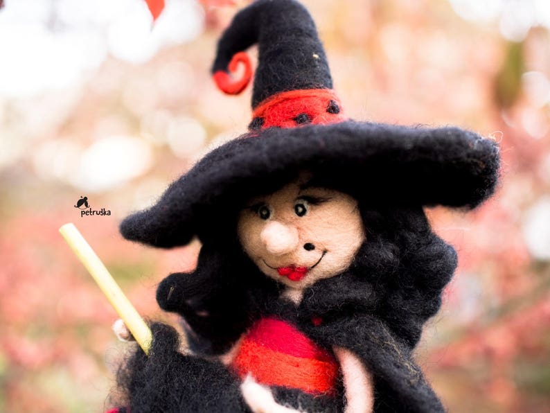 May include: A felt witch doll with a black hat and red and black dress. The doll is holding a yellow stick.