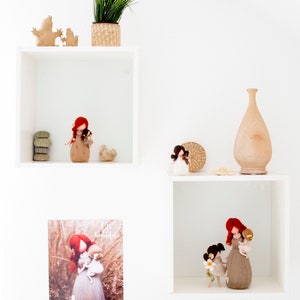 May include: White shelves with a variety of items, including a potted plant, a vase, and felt dolls. The dolls are all wearing brown dresses and have red hair. One doll is holding a smaller doll. The shelves are against a white wall.