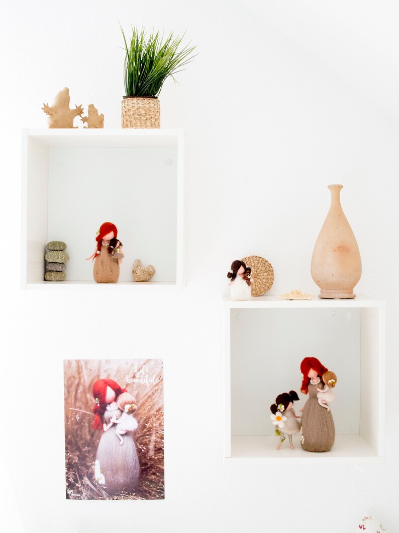 May include: White shelves with a variety of items, including a potted plant, a vase, and felt dolls. The dolls are all wearing brown dresses and have red hair. One doll is holding a smaller doll. The shelves are against a white wall.