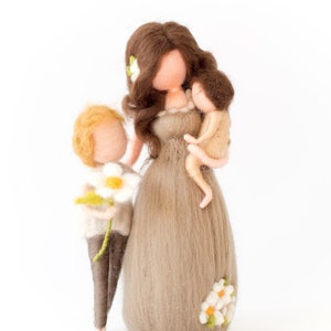 May include: A felt doll family with a mother holding a baby doll and a child holding a white flower. The mother doll has brown hair and is wearing a brown dress with white flowers. The child doll has blonde hair and is wearing brown pants and a white sweater.