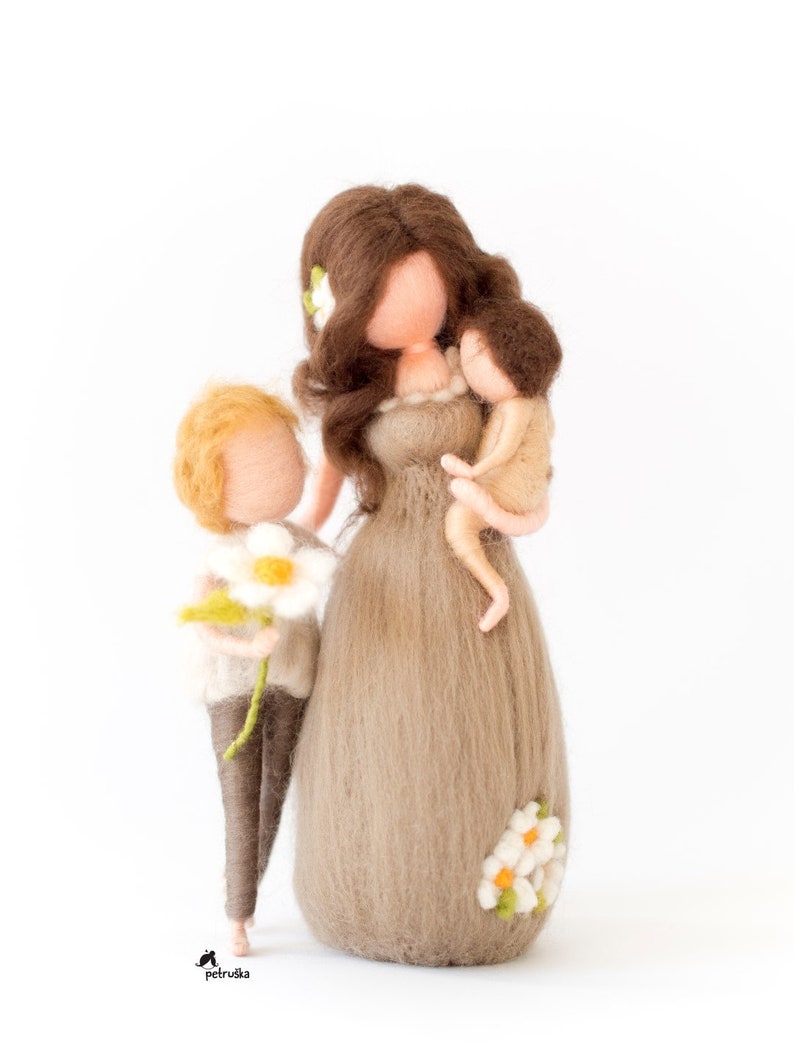 May include: A felt doll family with a mother holding a baby doll and a child holding a white flower. The mother doll has brown hair and is wearing a brown dress with white flowers. The child doll has blonde hair and is wearing brown pants and a white sweater.