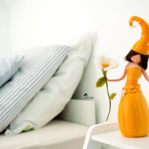 Peut inclure: Une poupée en feutre vêtue d'une robe et d'un chapeau jaunes, tenant une fleur blanche. La poupée est assise sur une étagère blanche avec un oreiller rayé blanc et bleu derrière elle.