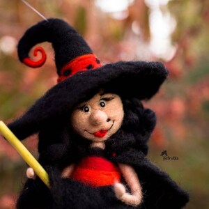 May include: A felt witch doll with black hair and a black hat with a red and black swirl. The doll is holding a yellow stick in her right hand and is wearing a red dress.