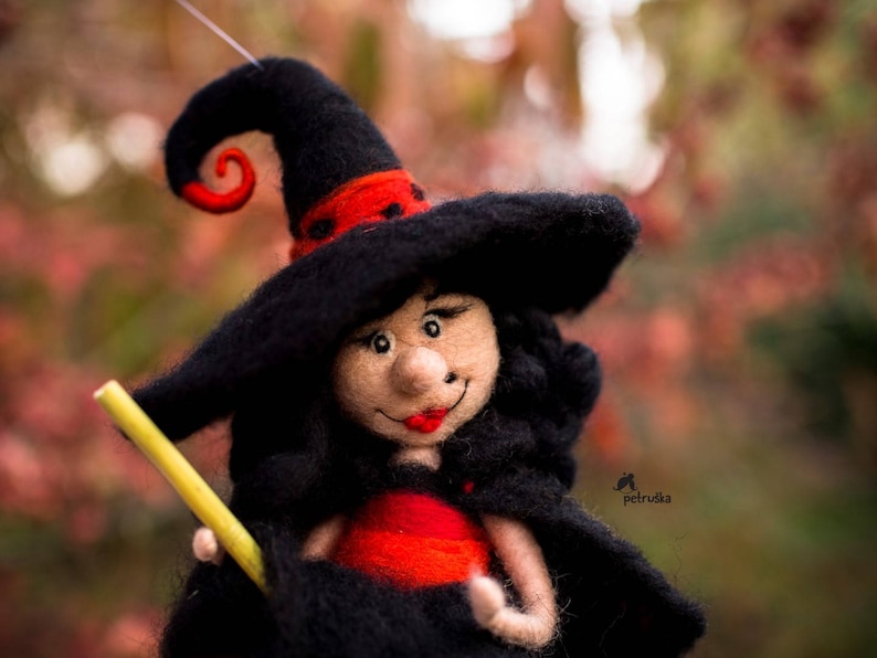 May include: A felt witch doll with black hair and a black hat with a red and black swirl. The doll is holding a yellow stick in her right hand and is wearing a red dress.