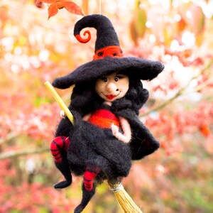 May include: A black felt witch doll with a red hat and red stockings is flying on a broom. The doll has a friendly face and is holding a stick. The background is a blurry image of fall foliage.