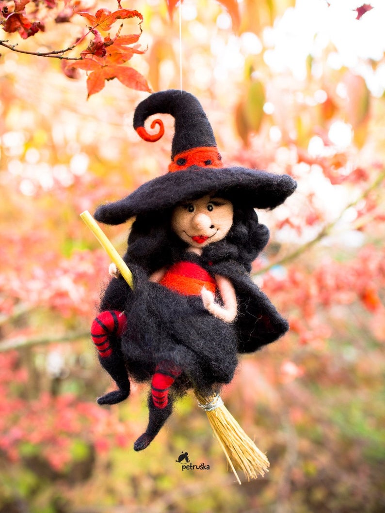 May include: A black felt witch doll with a red hat and red stockings is flying on a broom. The doll has a friendly face and is holding a stick. The background is a blurry image of fall foliage.