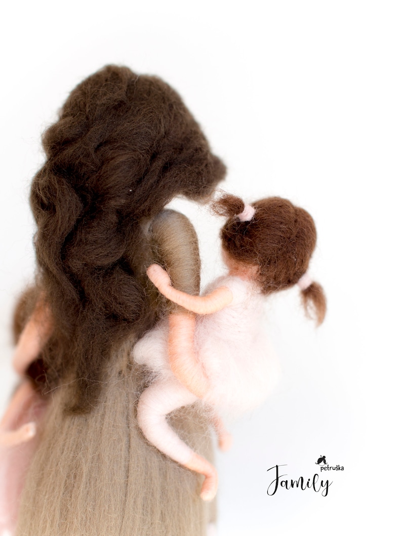 May include: A felt doll of a mother holding a smaller felt doll of a child. The mother has long brown hair and is wearing a light brown dress. The child has brown hair and is wearing a pink dress. The dolls are standing on a white background. The text "Family" is written in black below the dolls.