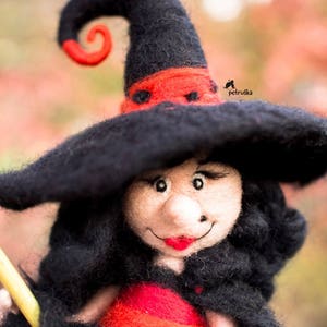 May include: A felt witch doll with a black hat and red trim, holding a broom. The doll has a smiling face with red lips and black eyes.